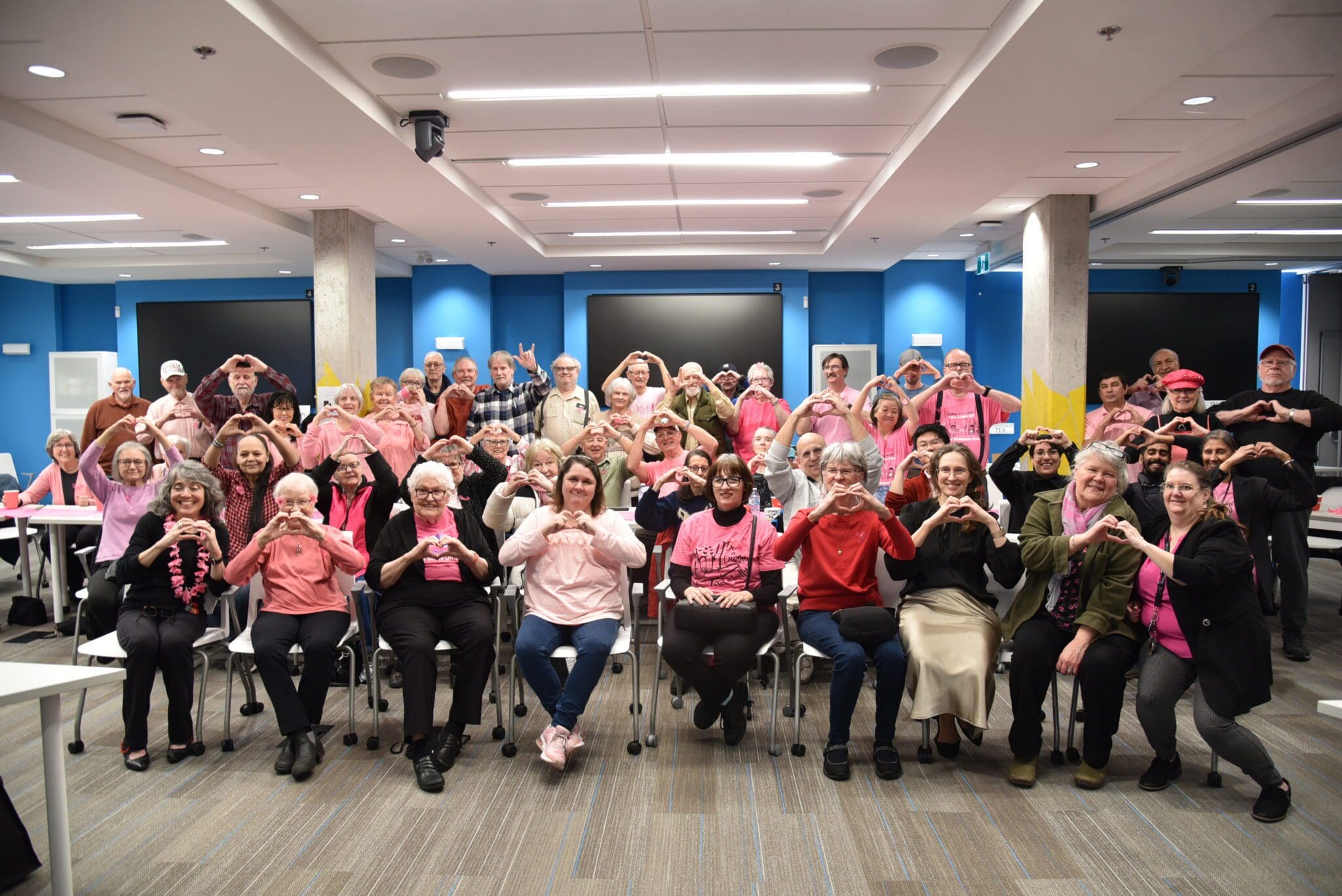 DSC_0339 A large group of Deaf, DeafBlind, and Hard of Hearing seniors gathered in a bright conference room at Wavefront Centre, wearing pink in support of Pink Shirt Day. They are smiling and making heart shapes with their hands, symbolizing kindness and inclusion. The room has blue walls, modern lighting, and tables arranged for a social event.